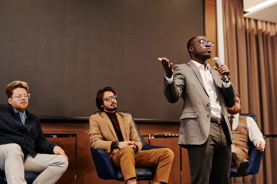 A diverse group of professionals engaged in a business panel discussion with a speaker presenting.